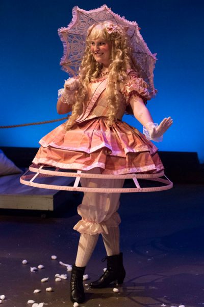 woman on stage with long blonde hair dressed in victorian modern style with a parasol and short dress showing stockings