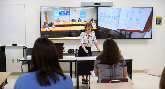 professor teaching a class with two screens behind her, one shows her book, the other shows the classroom on another campus.