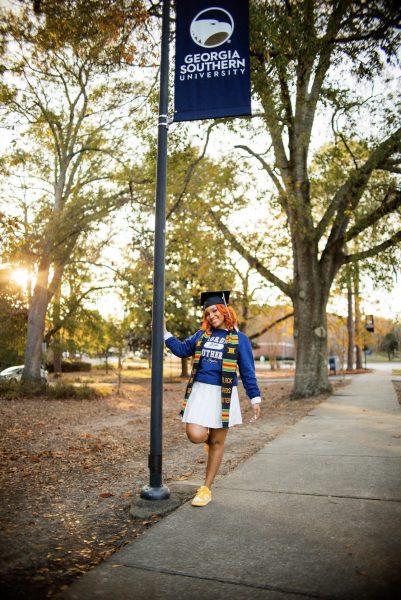 Taylor Pledger in a white skirt, blue Georgia Southern sweatshirt, yellow sneakers, graduation cap and stole. She is holding a pole with a blue Georgia Southern banner.
Photo Credit: Gregory Martin